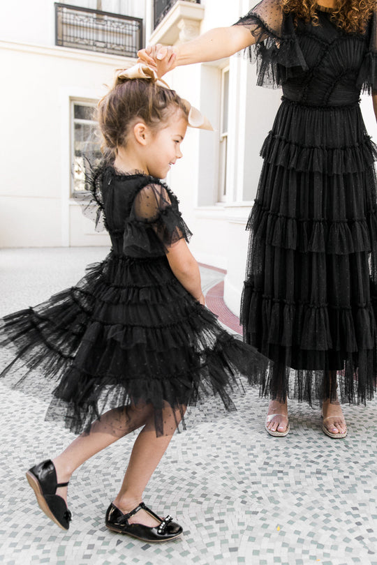 A young girl wears the Ivy City Co Mini Whimsical Dress in Black, featuring a layered tulle skirt and flutter sleeves, as she dances hand-in-hand with an adult in a coordinating black dress. Both smile brightly indoors atop a tiled floor.