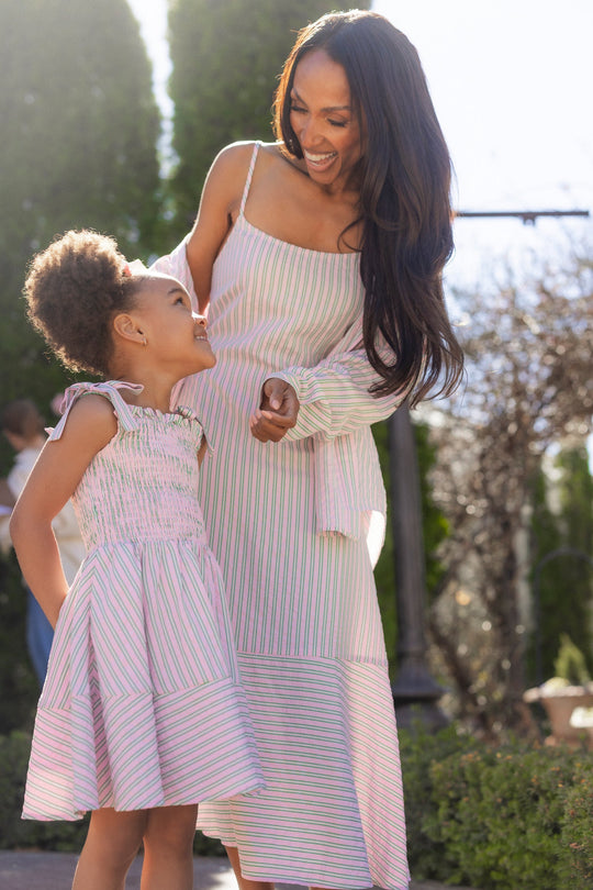 A woman and a young girl, both beaming at each other outdoors on a sunny day surrounded by lush greenery—the woman dressed in Ivy City Co’s Marbella Dress in Green & Pink Pinstripe, and the girl in a charming pink seersucker dress.
