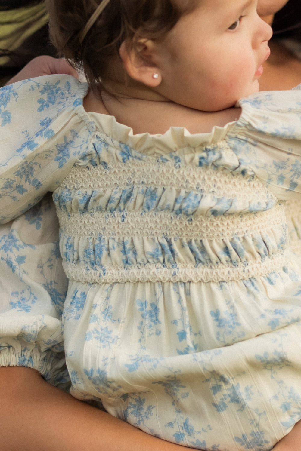 An infant dressed in the Ivy City Co Baby Roselyn Romper in Ivory Porcelain Fleur, featuring delicate blue floral patterns and charming ruffled details, sits sideways on someone’s lap.