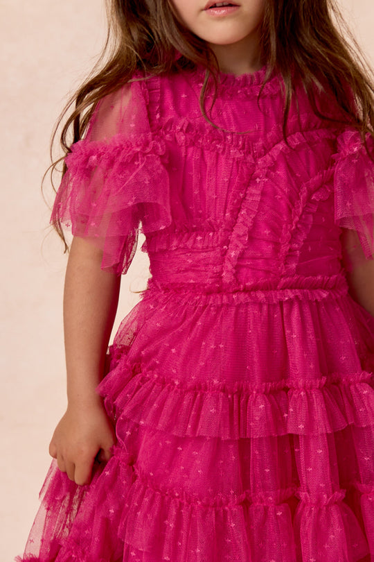 A young girl stands against a light background, wearing the Ivy City Co Mini Whimsical Dress in Fuchsia. The dress features a layered tulle skirt and sheer, flutter sleeves. Her long brown hair is loose, and her face is partially visible.