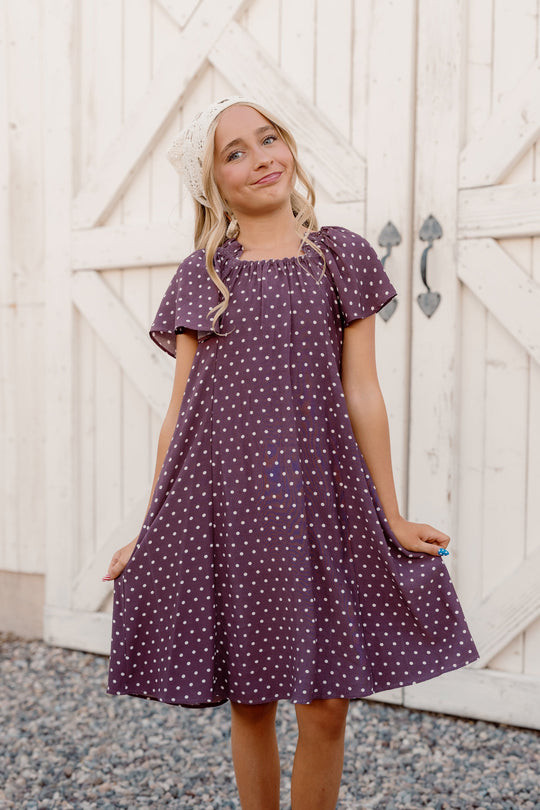 A young girl wearing the Ivy City Co Teen Blakely Dress in Aubergine Ditsy Dot, featuring flutter sleeves, smiles as she stands on gravel in front of white barn doors, holding the edges of her midi-length dress and sporting a white headscarf.