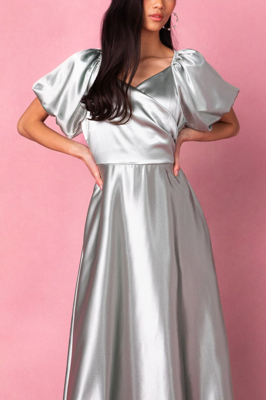 A woman in the Ivy City Co Tessie Dress in Sage poses against a pink background; her hands rest on her hips and her long dark hair falls in loose waves, embodying the graceful elegance of this modest bridesmaid style.
