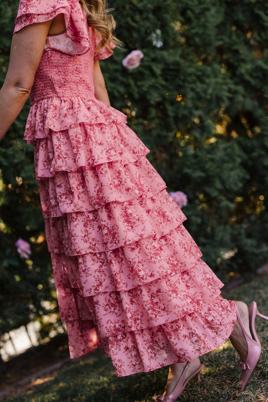 Clad in Ivy City Co’s Grace Dress in Desert Heritage Rose—a tiered, pink floral maxi featuring flutter sleeves—a person stands outdoors on the grass, surrounded by lush greenery and blooming pink flowers. Styled with pink heels, this maternity-friendly look embodies effortless elegance.