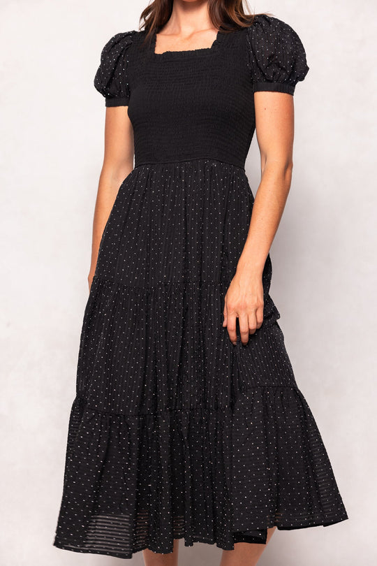 A woman stands against a light background wearing the Darby Dress in Black Diamonte by Ivy City Co—a black, short-sleeve, tiered midi dress featuring a smocked bodice, puff sleeves, and a flowy skirt adorned with small white polka dots. This style is effortlessly chic and maternity friendly.