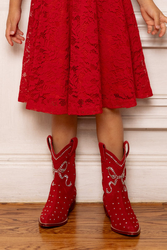 A child wearing a red lace dress and the Ivy City Co Mini Channing Bootie in Red stands on a wooden floor in front of a white wall—perfect for adorable mommy-and-me boots moments.