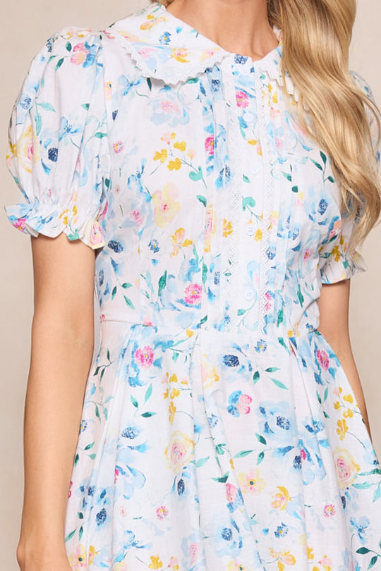 A woman wearing the Sophia Dress in Blue Bouquet by Ivy City Co, featuring a pastel floral print, puffed short sleeves, a wide collar, and lace trim, stands against a neutral background.