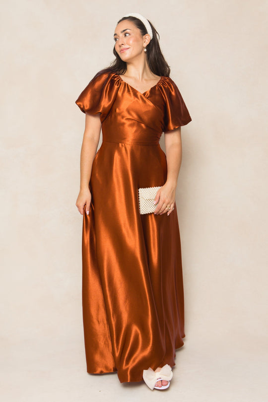 Wearing the Ivy City Co Tessie Dress in Amber Satin, this woman pairs the nursing-friendly gown with a white headband, white sandals, and a textured cream clutch—creating an elegant look that stands out perfectly for fall bridesmaids against a beige background.