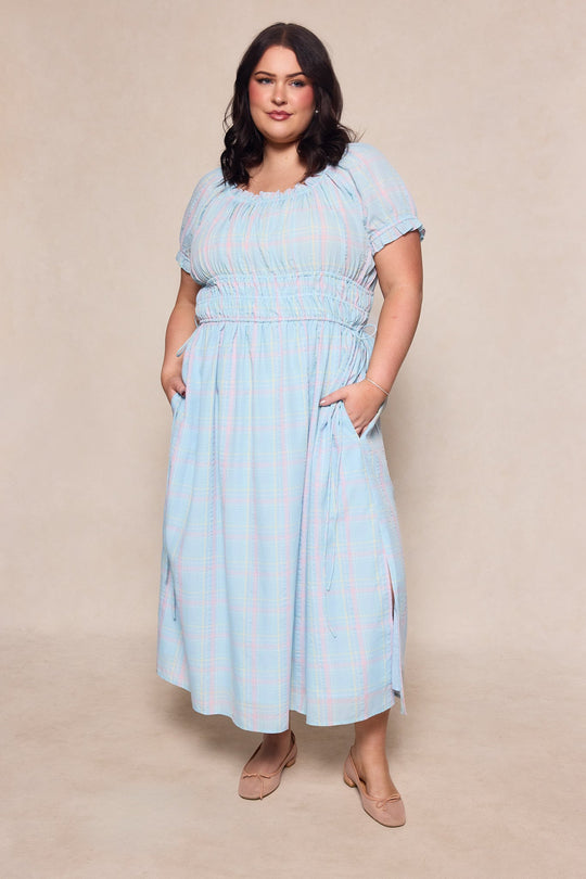 A woman with shoulder-length dark hair poses in front of a neutral background, wearing the Thalia Dress in Pastel Plaid by Ivy City Co. This stylish spring maxi dress features short sleeves and convenient pockets, offering a maternity-friendly fit that makes it perfect for any occasion. She completes her look with nude flats.