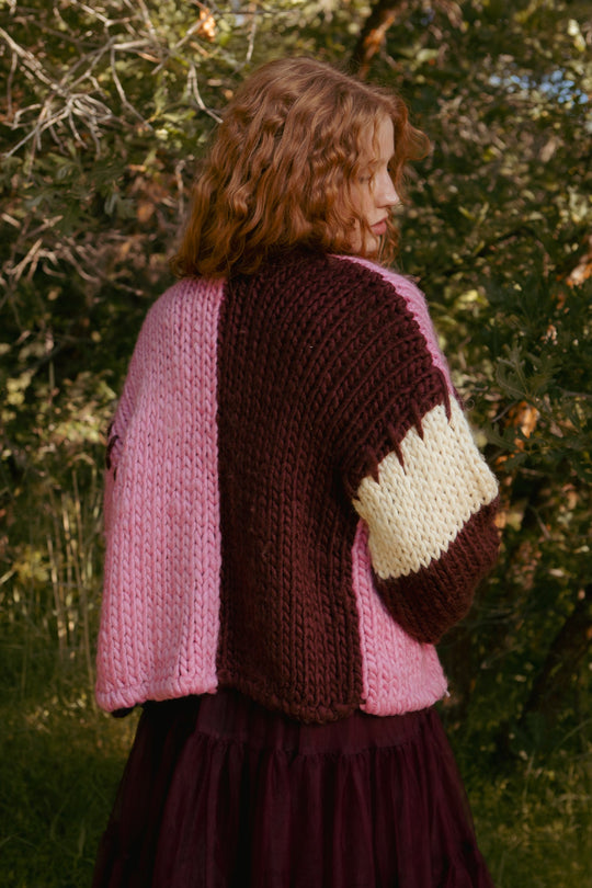 A person with wavy hair faces away, wearing the Ivy City Co Marissa Sweater in Pink & Red. They stand outdoors, surrounded by green trees and sunlight.