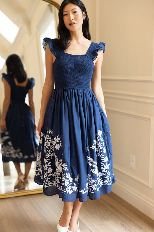 A woman wearing the Finch Dress in Blue by Ivy City Co stands indoors. She poses near a floor mirror, displaying both the front and back of her dress, which features white embroidery. The room has light-colored walls and wooden flooring.
