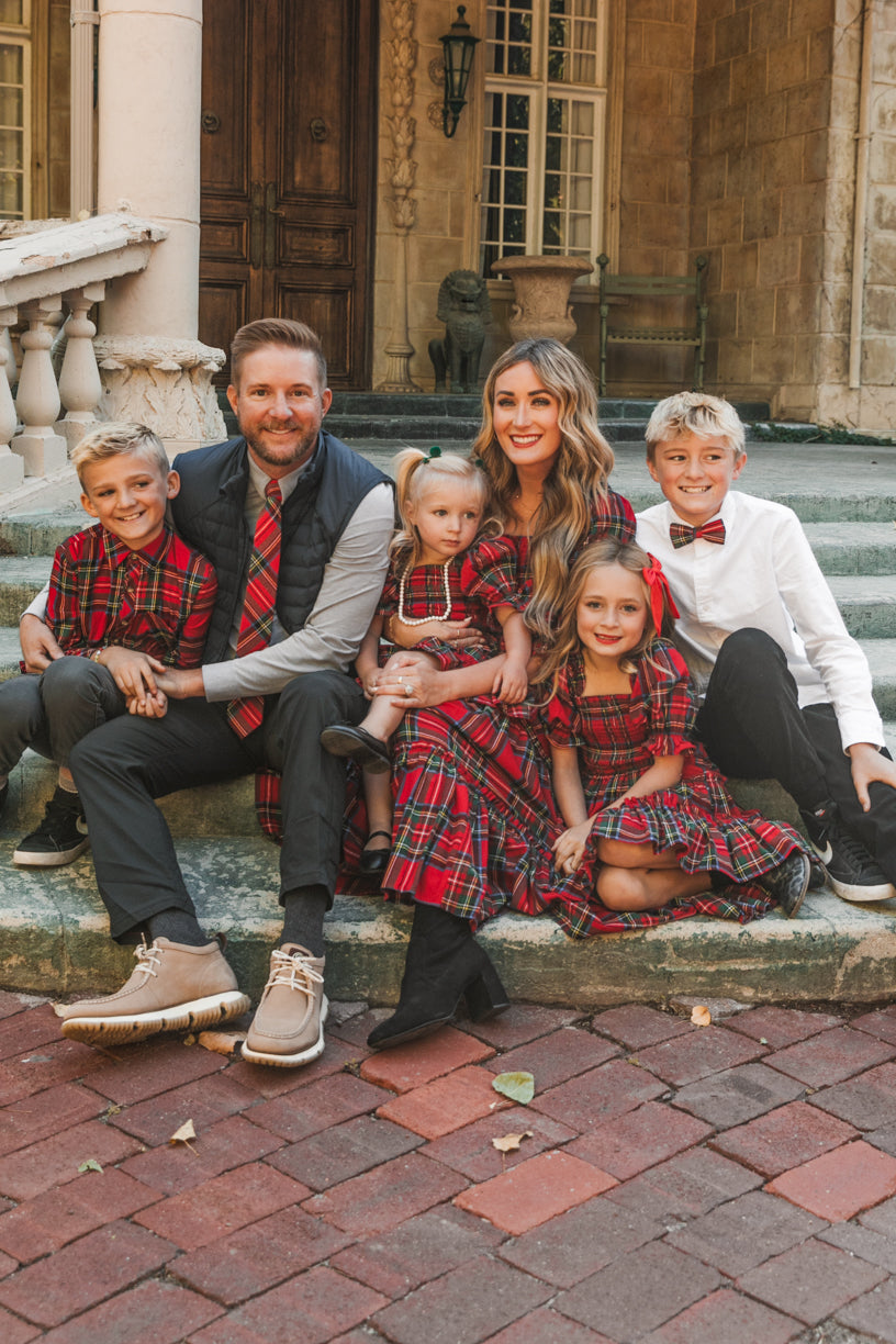 A family of six sits on stone steps, all smiling. The father, mother, three daughters, and two sons wear coordinated Holiday plaid outfits. They are in front of an old, elegant building with large windows and columns. The mother is wearing the Madeline Dress in Holiday Plaid by Ivy City Co—a maternity-friendly and nursing-friendly style that offers both comfort and festive flair.