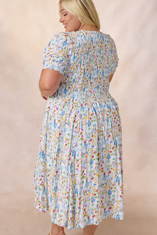 A woman with blonde hair stands against a soft beige background, wearing the Amelie Dress in Floral Fields by Ivy City Co—a light blue, floral midi dress featuring short sleeves and a smocked bodice gathered at the waist.