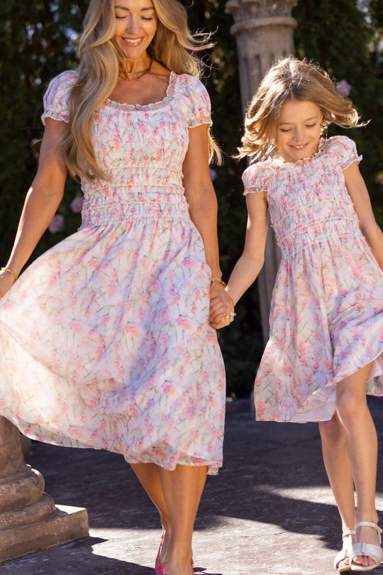 A woman and a girl, both wearing matching Kyla Dress in Watercolor Orchids by Ivy City Co, happily hold hands and twirl outdoors on a sunny day.