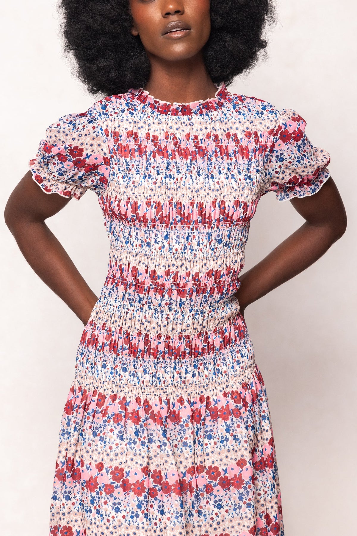 A woman wears the Ivy City Co Hayden Dress in Lush Garden—a short-sleeved, smocked floral midi dress with a red, pink, and blue pattern—standing with hands on hips against a plain background. This style is also maternity friendly.