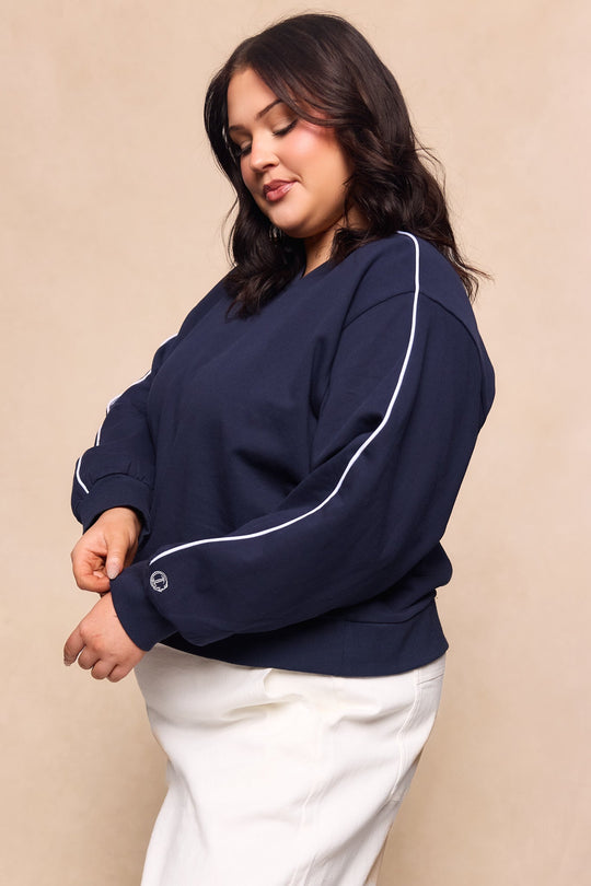 A woman with dark, wavy hair wears the Ivy City Co Kinsley Sweatshirt in Navy, featuring white piping on the sleeves, paired with a white skirt as she stands against a beige background.