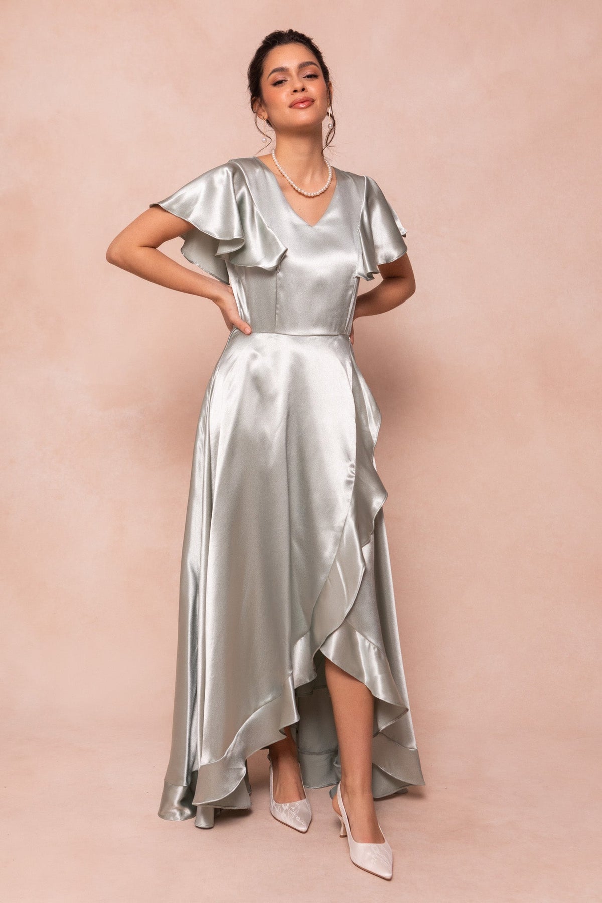 A woman poses against a peach background, smiling with her hands on her hips in the Callie Dress in Sage Satin by Ivy City Co—featuring shiny sage satin fabric, ruffled short sleeves, and an asymmetrical hem for a chic bridesmaid look.