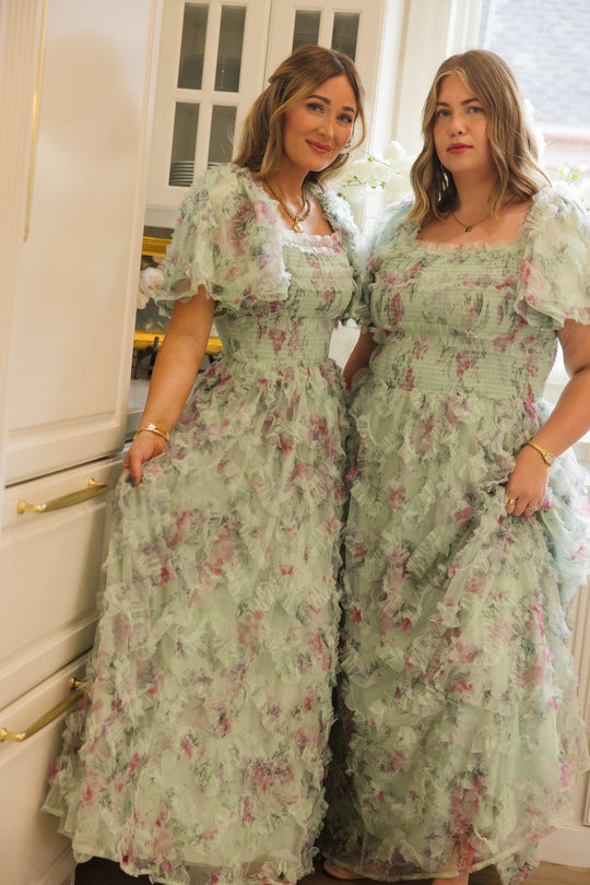 Two women stand together in a kitchen, both wearing matching Ivy City Co Florentina Dress in Pistachio Aquarelle Fleur. The soft chiffon dresses feature light green ruffles and a delicate floral pattern. Smiling gently, they rest their hands on the nursing friendly and maternity friendly dresses—an ideal choice for mothers. White cabinets and a window appear in the background.