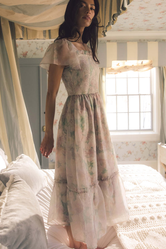 A woman in the Rhiannon Dress in Painted Bloom by Ivy City Co stands barefoot on a bed in a softly lit bedroom adorned with pastel decor and a canopy overhead. This delicate, maternity-friendly dress beautifully enhances the dreamy ambiance.