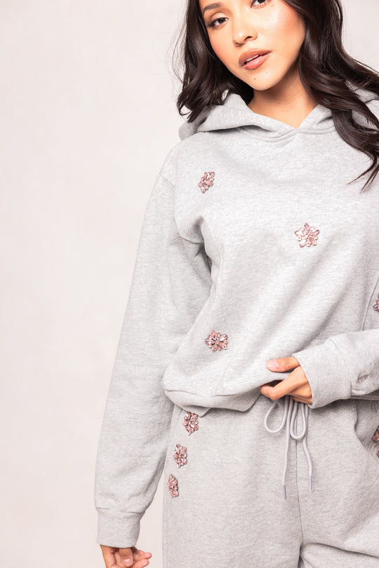 A woman stands against a plain light background wearing the Ivy City Co Skylar Hoodie in Heather Gray and matching sweatpants, both adorned with small pink floral designs. Her right hand rests on the soft fleece hoodie near her waist.