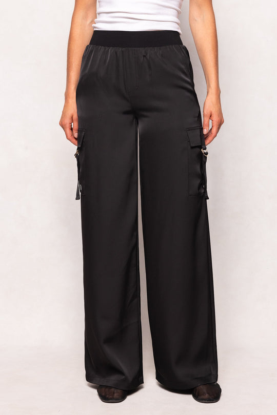 A person wearing the Ivy City Co Shay Satin Pants in Black, featuring a loose fit, elastic waistband, and side pockets, paired with a white top. Only the lower torso and legs are visible.