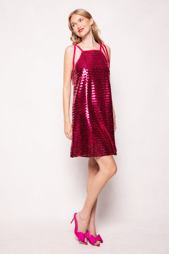 A woman wearing the Zelda Dress in Fuchsia Shingle Sequin by Ivy City Co, paired with matching pink heels, stands against a light background, smiling and looking to the side.