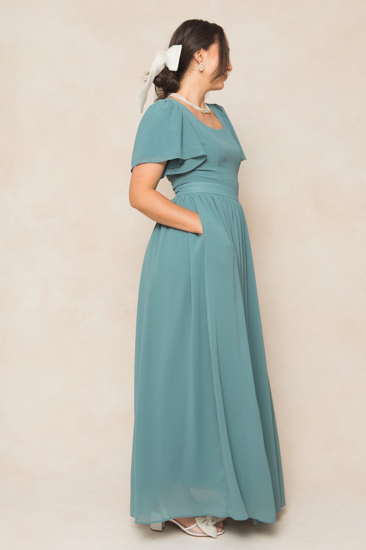 A woman stands sideways in the Ivy City Co Abbie Dress in Seaglass Chiffon—a teal, short-sleeve, floor-length bridesmaid gown with pockets. Styled with white heeled sandals, a pearl necklace, and a white hair ribbon, she embodies elegant ease thanks to the soft chiffon fabric. The look is set against a neutral beige background.