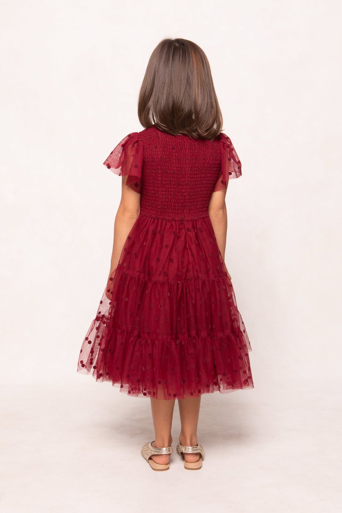 A young girl with brown hair stands facing away, wearing the Ivy City Co Mini Solange Dress in Burgundy Tulle, featuring short sleeves, ruffles, and polka dots, paired with beige sandals against a plain light background.