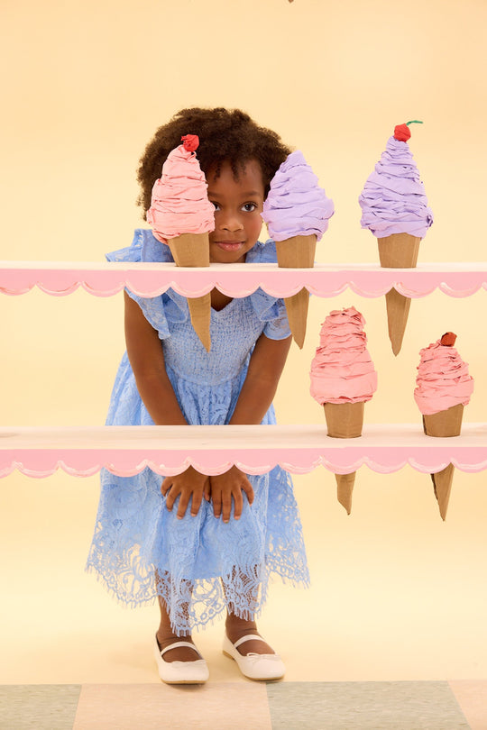 A young girl in the Ivy City Co Mini Hattie Dress in Cornflower Blue Lace pairs her look with white shoes as she crouches behind two pink shelves adorned with large, colorful ice cream cone props, set against a light yellow background—a perfect choice for playful mommy-and-me matching moments.
