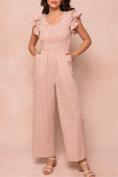 A woman wears the Ivy City Co Hattie Jumpsuit in Multicolor Gingham, featuring a smocked bodice, ruffled straps, wide legs, and side pockets; paired with scalloped white sandals. The background is light pink.