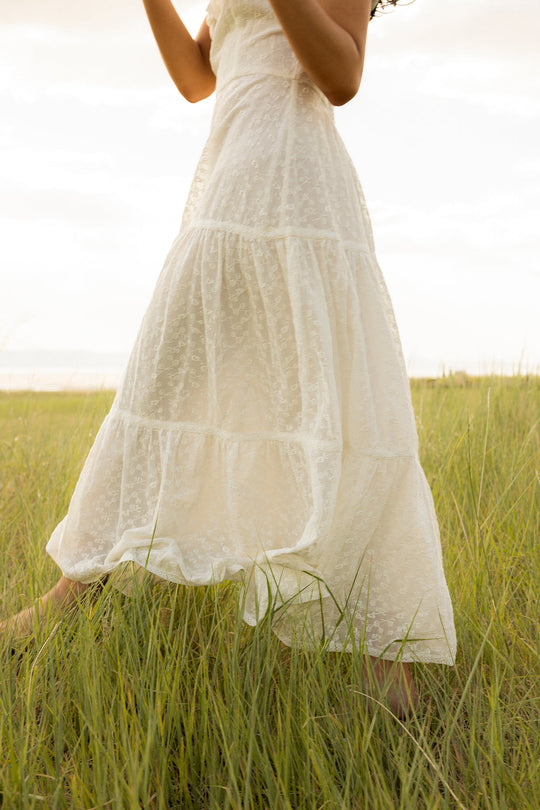 A woman, visible from her lower face and body, walks barefoot through tall green grass in a sunlit field wearing the Alaina Dress in Ivory by Ivy City Co—a white, sleeveless dress with a textured finish and smocked bodice.