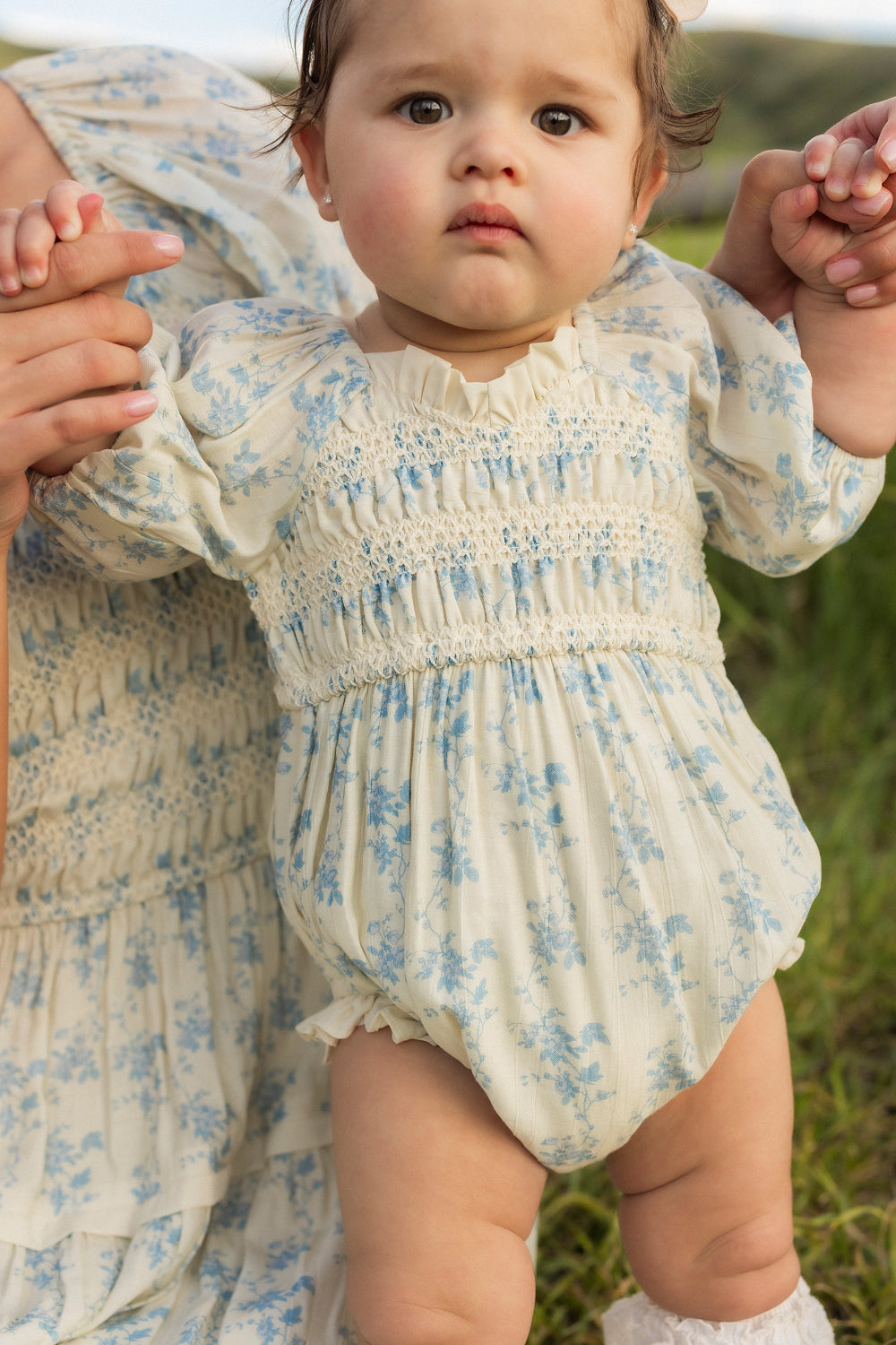 A baby wearing the Ivy City Co Baby Roselyn Romper in Ivory Porcelain Fleur stands outdoors, holding an adult’s hands for support. The romper features a delicate smocked bodice and matches the dress visible nearby. The baby gazes slightly upward with a neutral expression, surrounded by grass in this charming scene.
