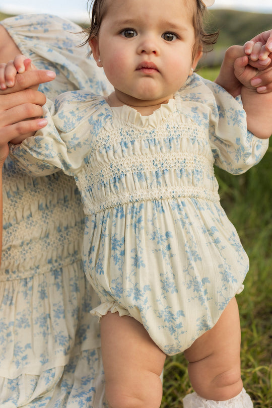A baby wearing the Ivy City Co Baby Roselyn Romper in Ivory Porcelain Fleur stands outdoors, holding an adult’s hands for support. The romper features a delicate smocked bodice and matches the dress visible nearby. The baby gazes slightly upward with a neutral expression, surrounded by grass in this charming scene.