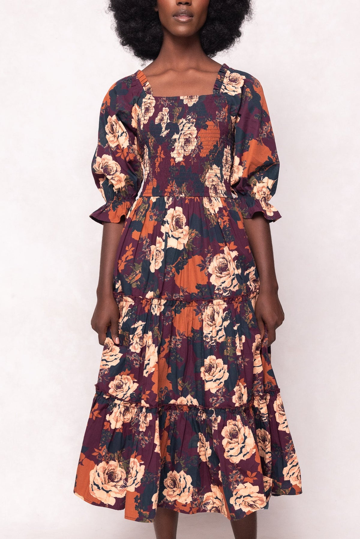 A woman wearing the Ivy City Co Madeline Dress in Equinox Rose, a maternity-friendly floral midi dress featuring puffed sleeves, a smocked bodice, and a tiered skirt in orange, burgundy, and cream tones with bold rose patterns.