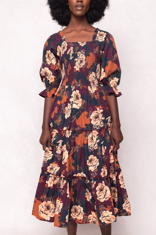 A woman wearing the Ivy City Co Madeline Dress in Equinox Rose, a maternity-friendly floral midi dress featuring puffed sleeves, a smocked bodice, and a tiered skirt in orange, burgundy, and cream tones with bold rose patterns.