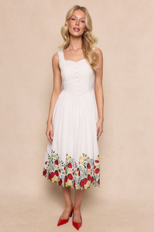 A woman stands against a beige background wearing the Ivy City Co Clarissa Dress in Strawberry, a white sleeveless dress featuring colorful floral embroidery at the hem, paired with red pointed heels.