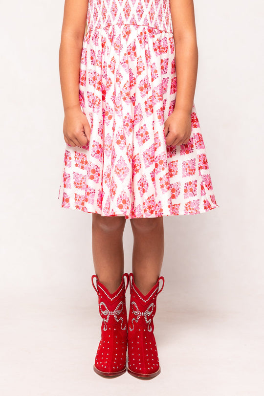 A child stands with arms by their sides, sporting the Ivy City Co Mini Channing Bootie in Red—a bright, playful cowboy-style boot adorned with white details—paired here with a white and pink dress featuring red floral patterns, perfect for a charming mini-me look that coordinates effortlessly with matching mommy-and-me boots.