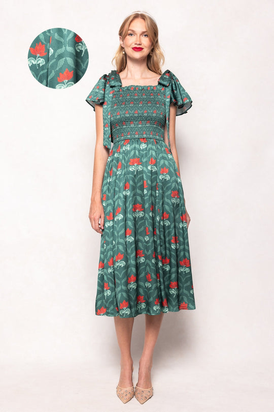 A woman models the Ivy City Co Rayna Dress in Green Floral Block—a maternity-friendly style with short ruffled sleeves, a smocked bodice for a flattering fit, and a red floral pattern on green. A close-up inset reveals the intricate fabric detail. She stands against a plain light background, wearing pointed beige shoes.