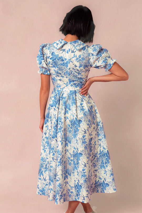 A woman with short dark hair is shown from the back against a plain light background wearing the Ivy City Co Sophia Dress in Blue Floral, featuring puffed sleeves and a fitted waist.