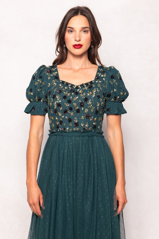 A woman wearing the Ivy City Co Adara Dress in Green—a dark green maxi dress featuring puff sleeves and floral embroidery—stands against a light background, looking at the camera with red lipstick and loose, wavy brown hair.