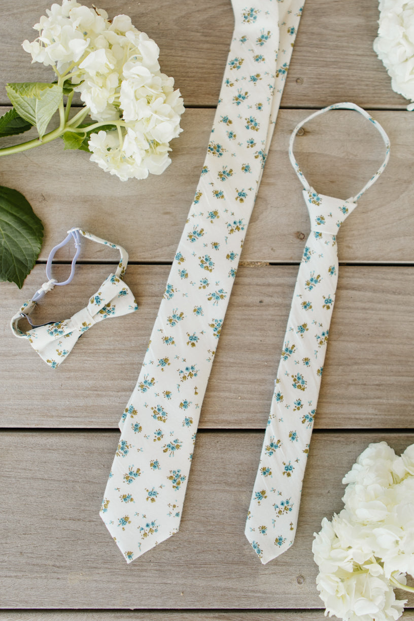 Three “Ties in Madeline Blue Floral” by Ivy City Co—a necktie, child’s tie, and boys’ bow tie—are displayed on a wooden surface surrounded by white hydrangea flowers, ideal for coordinating stylish family matching outfits.
