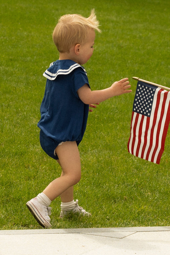 A young child wearing the Ivy City Co Baby Sailor Romper in Navy and white shoes walks on grass, reaching toward a small American flag.