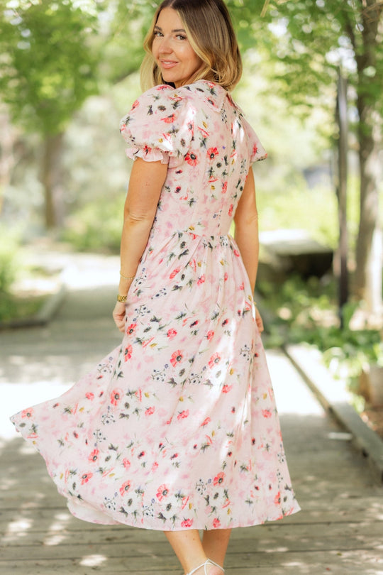 A woman stands on a wooden path outdoors, looking over her shoulder and smiling as sunlight filters through lush green trees. She wears the Sophia Dress in Roselyn Pink by Ivy City Co, its pink floral design perfectly matching the soft, summery atmosphere.