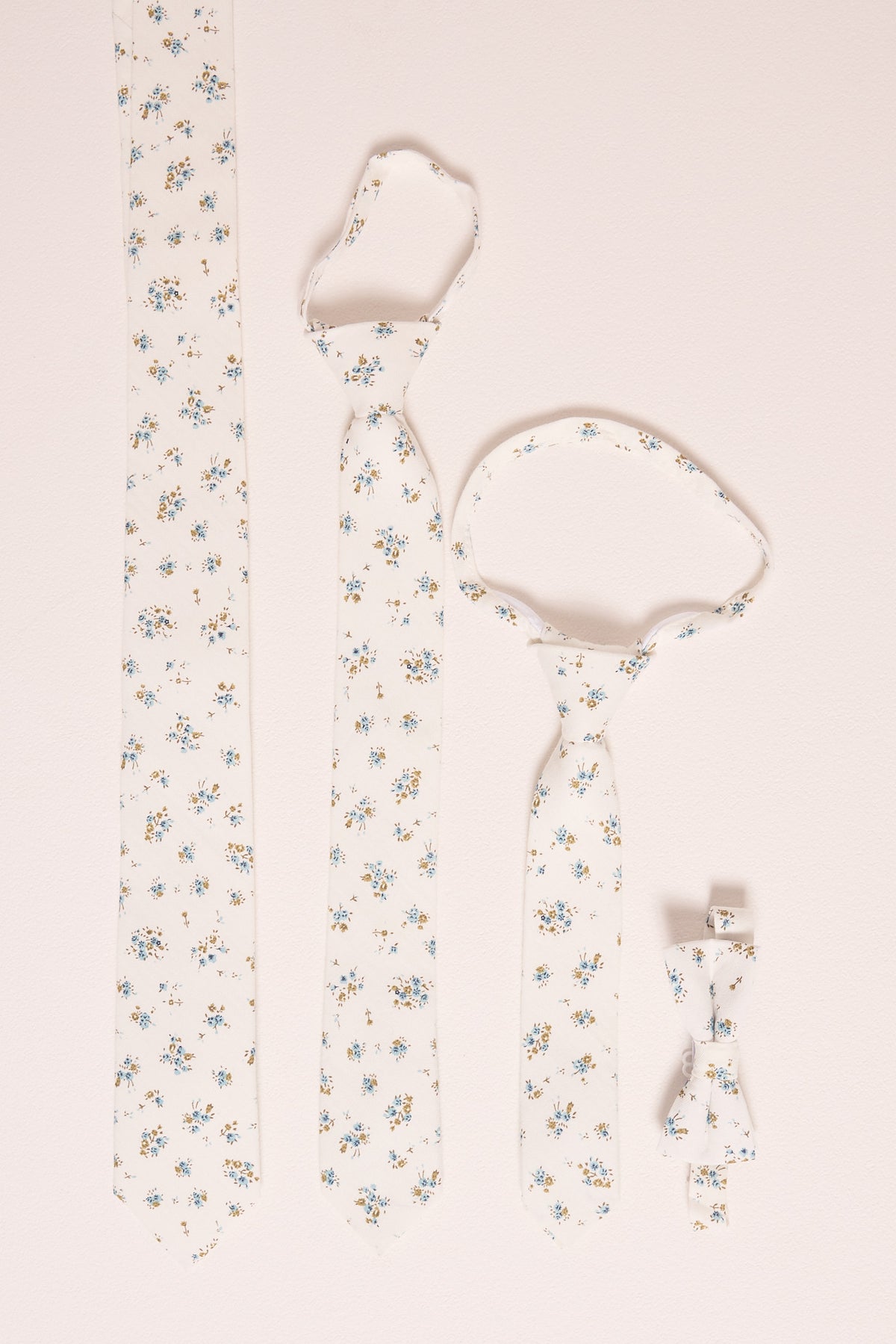 Four Ivy City Co 'Ties in Madeline Blue Floral' are displayed on a light surface: adult, child, and baby ties, plus a boys' bow tie—all featuring delicate blue and yellow floral patterns for perfect family matching.