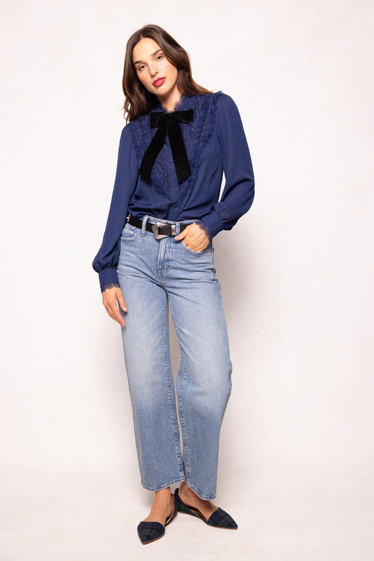 A woman with long dark hair stands against a white background wearing the Ivy City Co Blythe Blouse in Navy, a nursing-friendly top, paired with light blue high-waisted jeans, a black belt, and black pointed flats. She poses with one hand in her pocket.