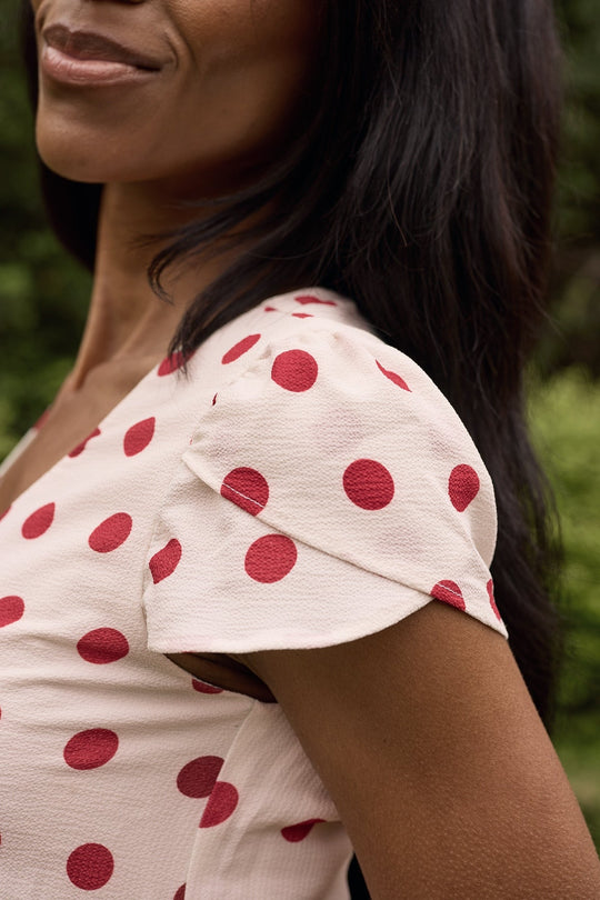A woman models the Ivy City Co Fiona Dress in Red Polka Dot outdoors, her lower face, shoulder, and upper arm visible as she offers a subtle smile.