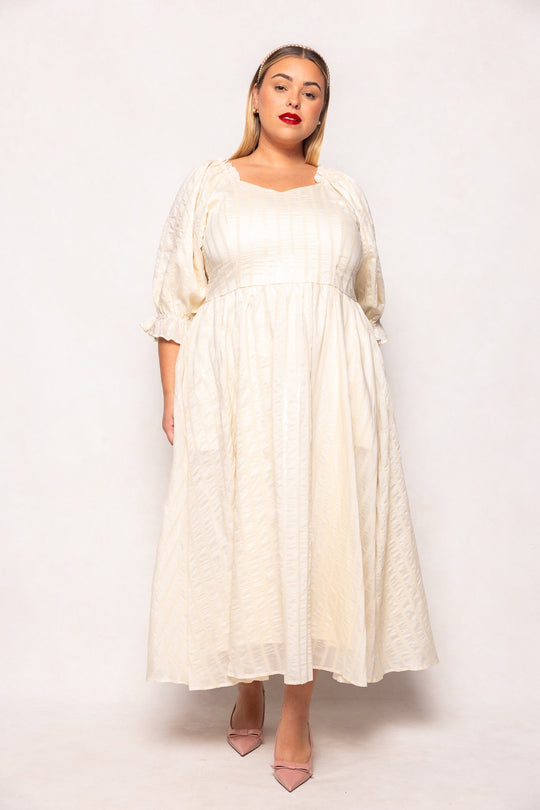 A woman wearing the Ivy City Co Tindra Dress in Cream Sequin, a stylish cream-colored midi dress with puffed sleeves that is both nursing-friendly and perfect for maternity wear, stands against a white background. She pairs the look with pink heels and wears her straight blonde hair loose, finishing the outfit with bold red lipstick.