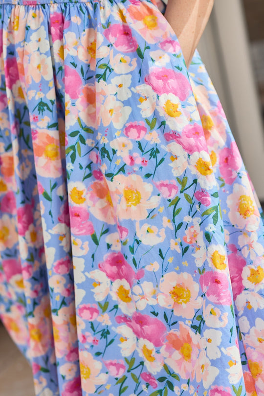A close-up of the Teagan Dress in Delia Blue by Ivy City Co—a blue smocked cotton dress featuring a vibrant floral pattern in pink, yellow, and white—shows part of a person’s arm and hand tucked into the dress pocket.