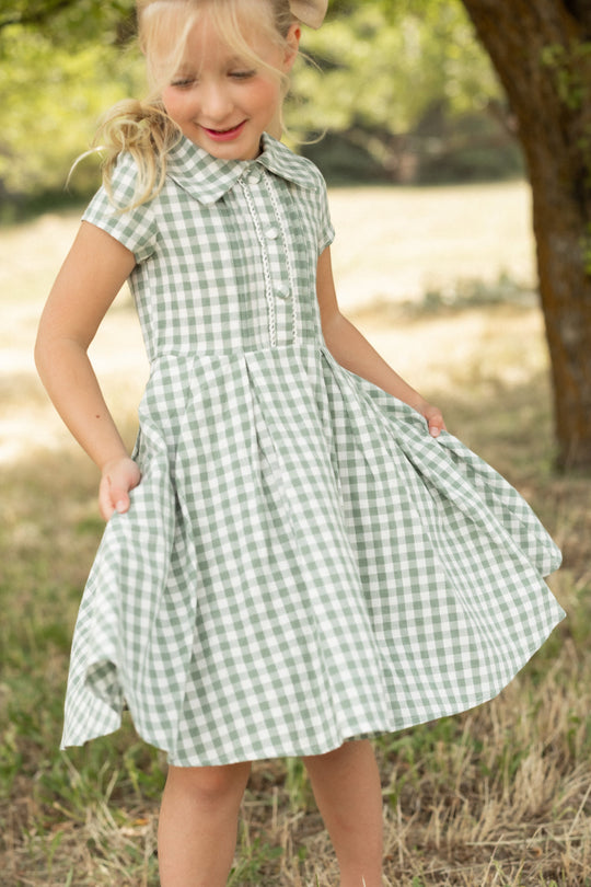 A young girl stands on the grass near a tree, smiling as she holds the sides of her pleated skirt while wearing the Ivy City Co Mini Amber Dress in Sage Gingham.