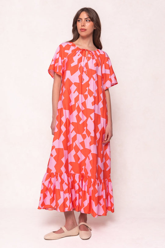 A woman wearing the Ivy City Co Haven Dress in Geometric—a loose, ankle-length dress with pink and orange geometric patterns, short sleeves, and a ruffled hem—paired with light-colored flat shoes, stands against a plain background.