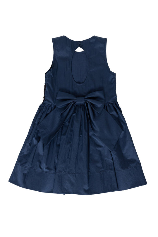 The Mini Greyson Dress in Navy by Ivy City Co is a charming sleeveless dress for children, featuring a large bow at the back and a round cutout near the neckline. Displayed on a plain white background, this stylish and comfortable dress is perfect for special occasions.